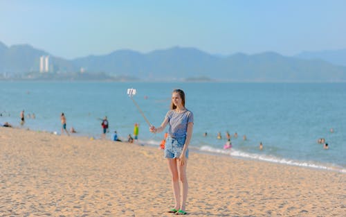 女人站在海边采取自拍照在白天使用独脚架 · 免费素材图片