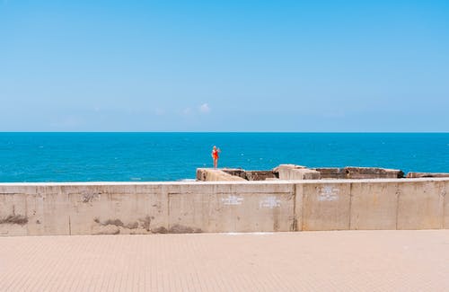 站在混凝土墙附近海水在湛蓝的天空下的人 · 免费素材图片