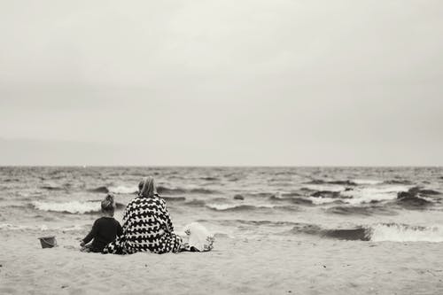 女人和孩子坐在水附近的沙滩上 · 免费素材图片