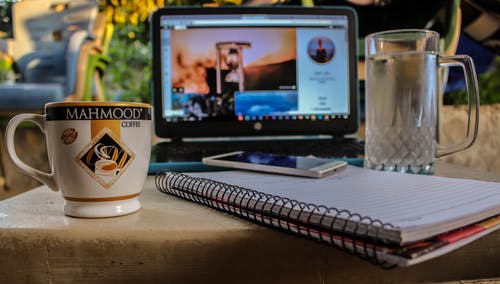 陶瓷杯和玻璃啤酒杯之间的螺旋笔记本上的白色智能手机附近黑色电脑显示器显示小时玻璃 · 免费素材图片