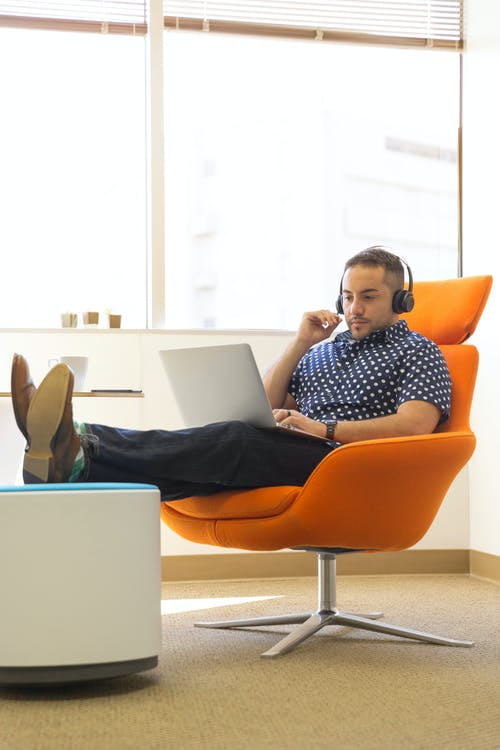 使用便携式计算机时坐在橙色软垫椅子上的男人戴着耳机 · 免费素材图片