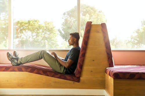 坐在玻璃窗口旁边使用笔记本电脑的褐红色和棕色织物填充沙发上的男人 · 免费素材图片