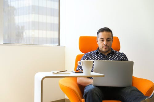 男子坐在橙色沙发椅上，他膝上的灰色便携式计算机 · 免费素材图片