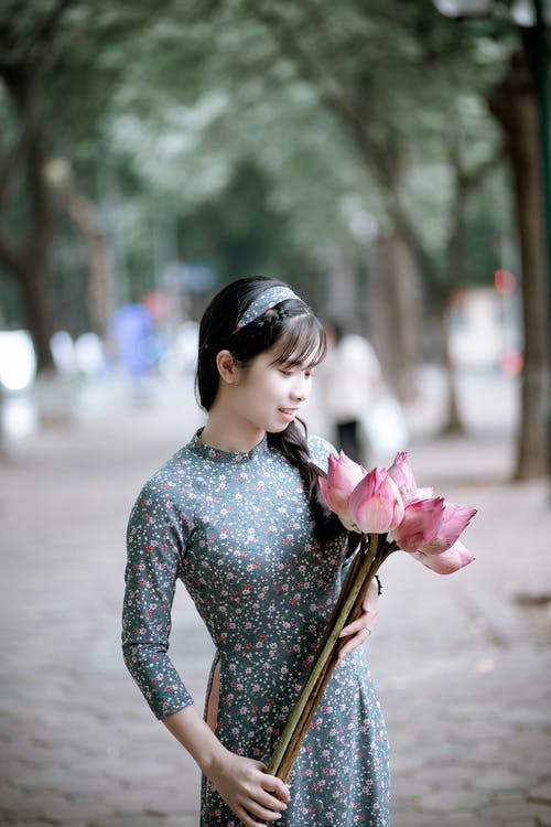 女人抱着粉红色莲花花束在绿树旁的路上 · 免费素材图片