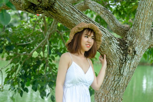戴着棕色帽子站在绿树下的女人 · 免费素材图片