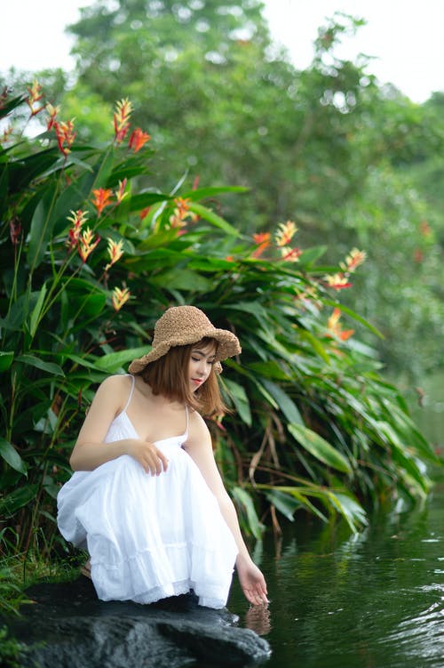 女人穿着白色连衣裙蹲在植物附近的水体附近 · 免费素材图片