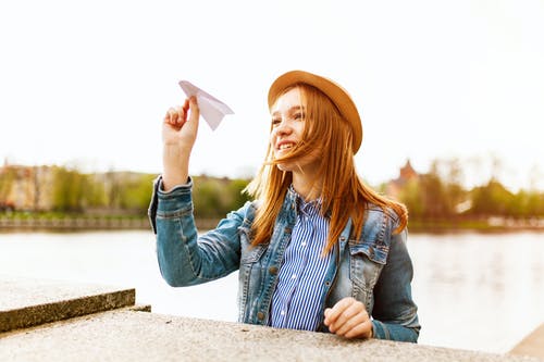蓝色牛仔纽扣式外套和棕色帽子的女人即将在水体旁边飞纸飞机 · 免费素材图片