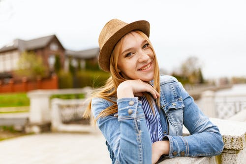 微笑的女人，穿着蓝色牛仔纽扣式外套和棕色的帽子，用手在她的脸上 · 免费素材图片