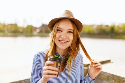 微笑的女人，穿着白色和蓝色的条纹的纽扣式上衣衬衫，拿着塑料咖啡杯站附近的水体 · 免费素材图片