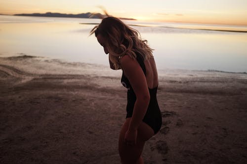 女人穿着黑色一件比基尼站在黄昏时在海滩附近的岸上 · 免费素材图片
