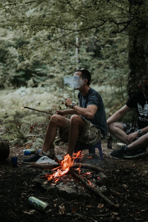 男子吸烟，坐在凳子上燃烧的木头附近 · 免费素材图片