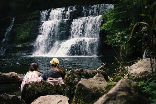 两个女人坐在瀑布附近的岩石上 · 免费素材图片