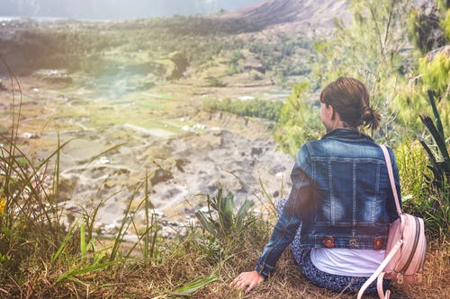女人穿着蓝色牛仔夹克，坐在悬崖上，俯瞰开放领域 · 免费素材图片