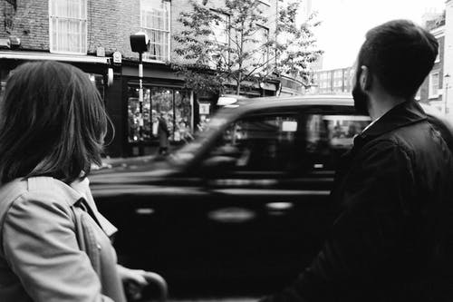 灰度摄影男人和女人站在建筑物附近道路上的汽车时 · 免费素材图片
