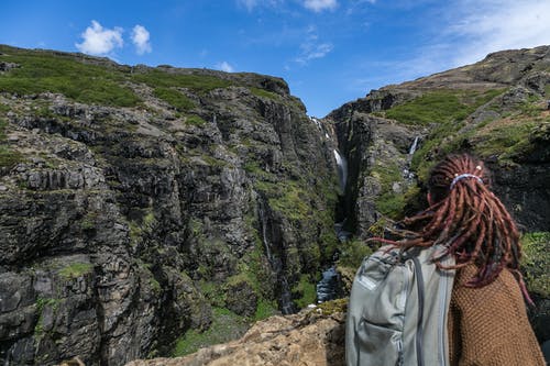 穿着棕色毛衣和灰色背包和棕色的头发辫子在山上俯瞰瀑布的人 · 免费素材图片