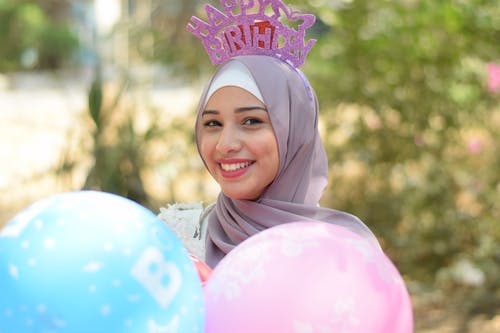 微笑的女人，戴着灰色的盖头头巾，拿着粉色和蓝色的气球 · 免费素材图片