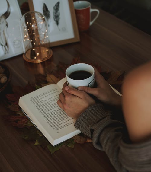 拿着杯子咖啡上面被打开的书的女孩在棕色木桌上 · 免费素材图片