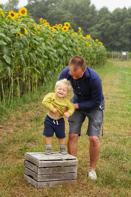 男人抱着微笑的孩子站在向日葵附近的棕色木箱上 · 免费素材图片