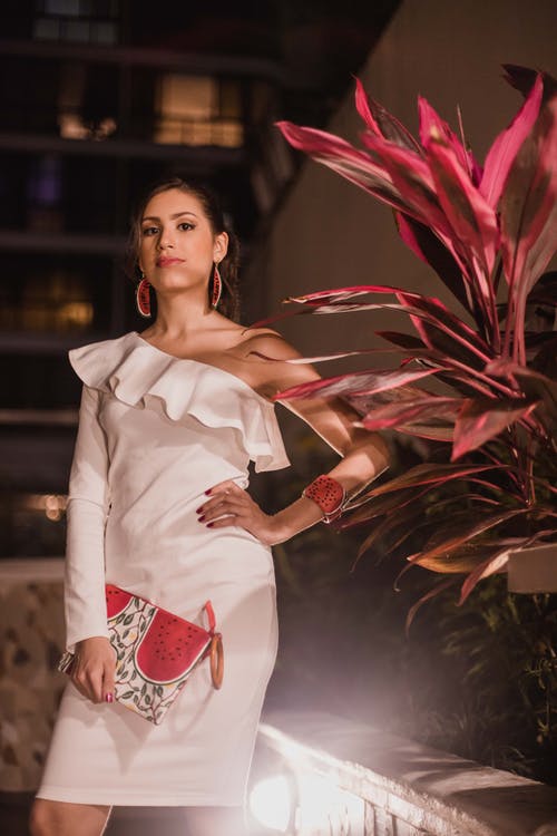 拿着袋子的白色礼服的妇女站立在植物旁边在夜间 · 免费素材图片