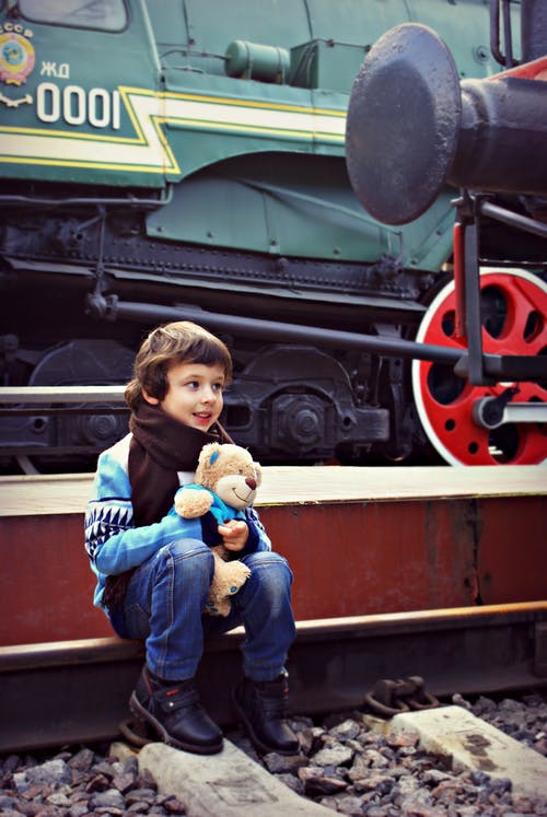 男孩抱着熊毛绒玩具坐在火车附近 · 免费素材图片