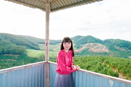 站立在篱芭旁边的女孩在白云和蓝天下的山附近 · 免费素材图片