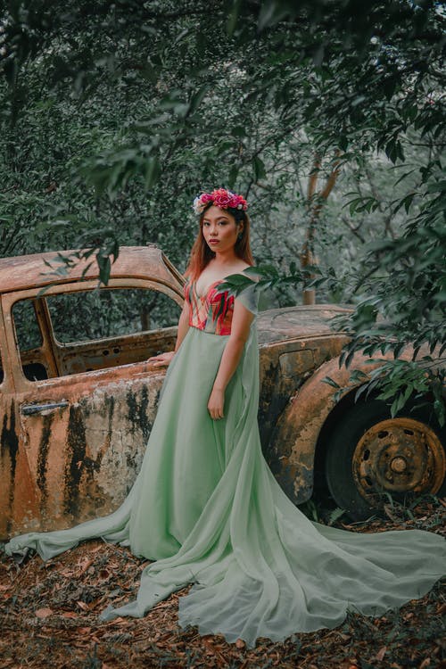 女孩穿着蓝绿色和红色礼服站在废弃的车辆附近 · 免费素材图片