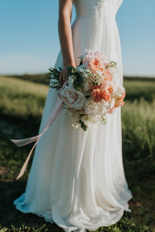 女人穿着白色婚纱，拿着花束站在绿色的原野上 · 免费素材图片