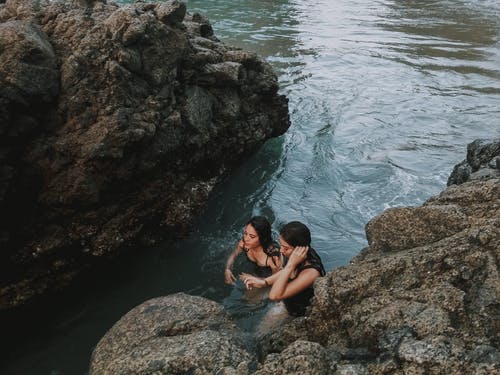 两名妇女在岩石峭壁旁边的水体上游泳 · 免费素材图片