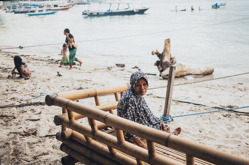 女人穿着黑色上衣，坐在海边的棕色竹凳上 · 免费素材图片
