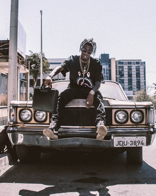 男子手持黑色袋子坐在汽车引擎盖上的照片 · 免费素材图片