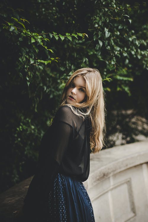 女人靠在绿色的树叶树附近的墙上 · 免费素材图片