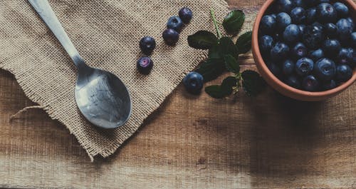 蓝莓在勺子附近的杯子和棕色表面上 · 免费素材图片