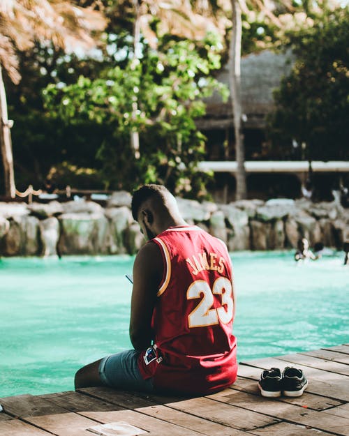 男子身穿红色勒布朗·詹姆斯23球衣坐在水体附近的码头上 · 免费素材图片