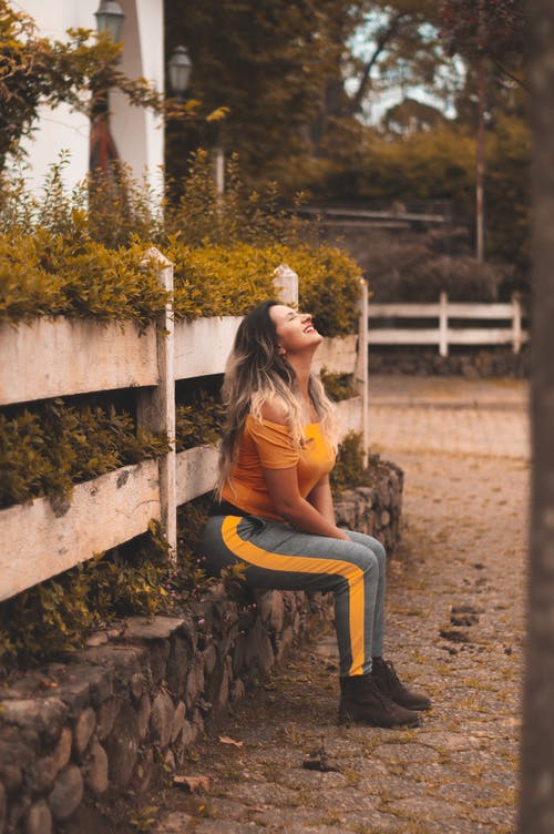 女人穿着橙色的露肩衬衫和灰色的裤子，坐在壁架上 · 免费素材图片