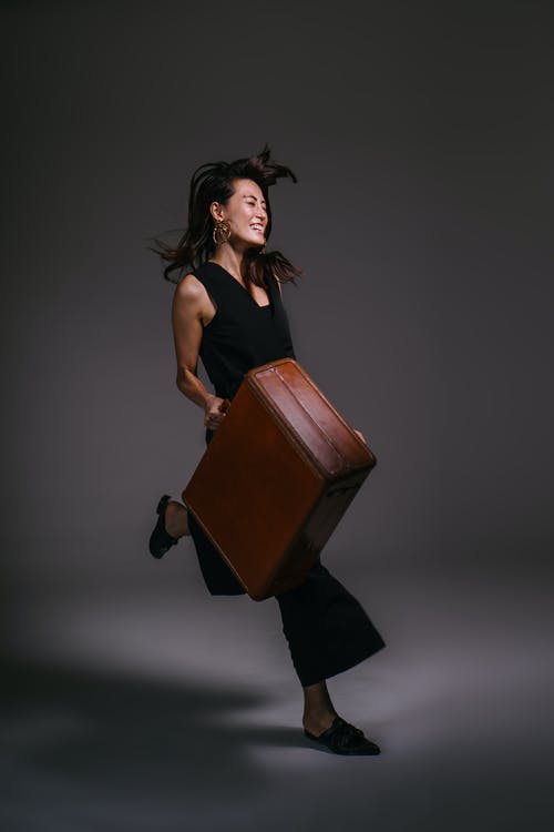 拿着棕色皮革手提箱的微笑的女人的照片 · 免费素材图片