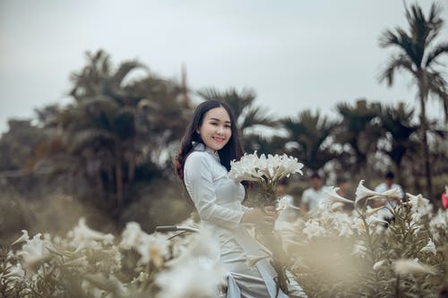 白色礼服的妇女站立和拿着白色瓣花和微笑 · 免费素材图片
