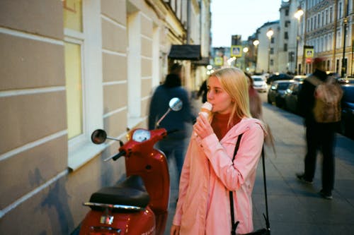 在人行道旁的红色小型摩托车附近吃冰淇淋的女人的照片 · 免费素材图片