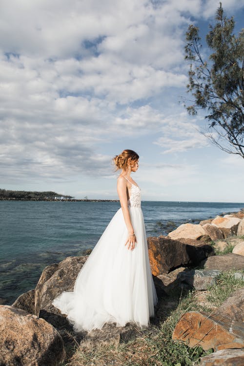站立在岩石上的妇女在水体附近穿白色婚礼礼服 · 免费素材图片