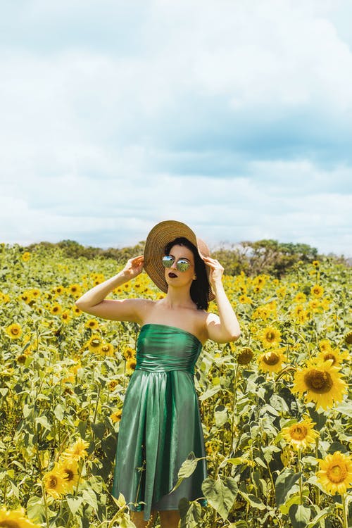 女人穿着绿色的管礼服和遮阳帽站在向日葵的草地中间 · 免费素材图片