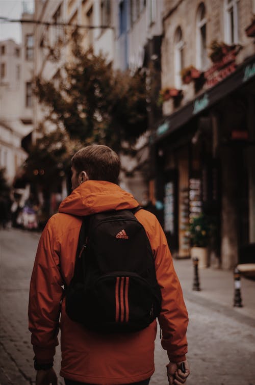 穿橙色夹克，黑色阿迪达斯背包的男人走在路中间 · 免费素材图片