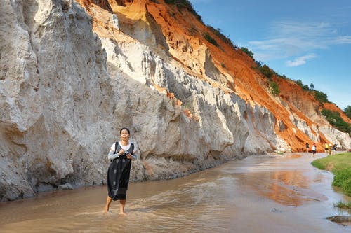 女人站在蓝色和白色的天空下的岩石山附近的水体 · 免费素材图片