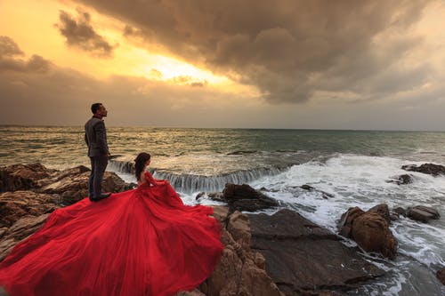 穿着红色礼服的女人和穿着灰色西装的男人站和坐在海边的巨石上 · 免费素材图片