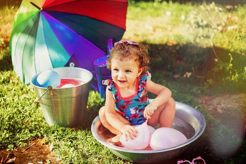 小女孩微笑着坐在灰色的不锈钢盆玩粉红色的气球 · 免费素材图片