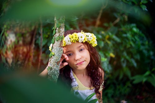 白色和黄色背心和黄色和白色花卉头饰倚在树干上的女孩 · 免费素材图片