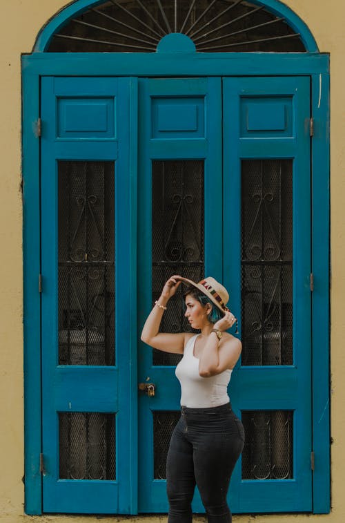 按住她的帽子站在蓝色的门附近的女人 · 免费素材图片
