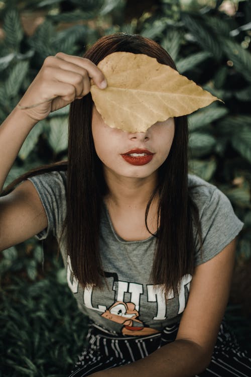 女人在灰色的t恤，捂着脸，用棕色的叶子的特写照片 · 免费素材图片