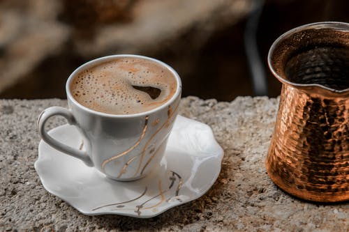 白色陶瓷杯碟与黄铜色的咖啡壶旁边的咖啡里面的特写照片 · 免费素材图片
