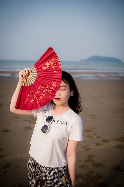 女人站在海边抱着风扇隐藏她的脸的一部分的照片 · 免费素材图片