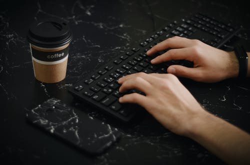 在电话和咖啡杯旁边使用键盘的人 · 免费素材图片