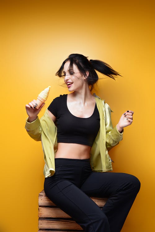 微笑的女人，穿着黑色活跃的衣服，坐在木箱在黄色背景前按住冰淇淋蛋筒的照片 · 免费素材图片